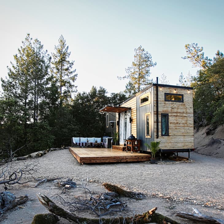 Tiny house with wooden siding on a gravel lot, surrounded by trees, featuring a small deck with outdoor seating.