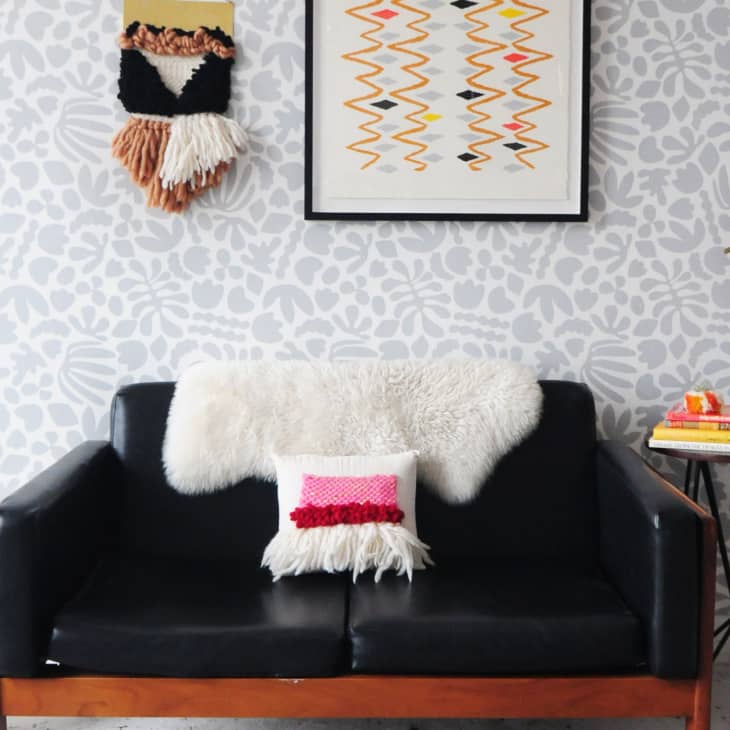 Black sofa with white fur throw, pink cushion, cactus, and side table with books against a patterned wall with woven art.