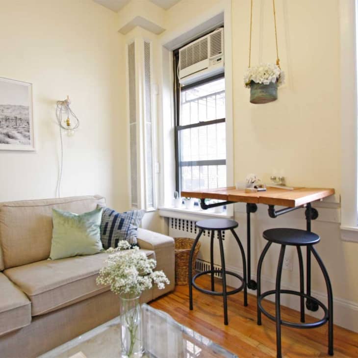 Cozy living room with beige sofa, green and blue pillows, wall art, and a small wooden table with two stools by the window.
