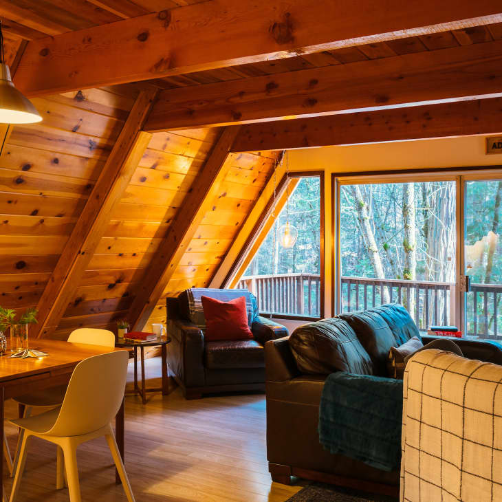 A-frame cabin interior with wooden walls, dining table, chairs, leather sofas, and large windows overlooking a forest.