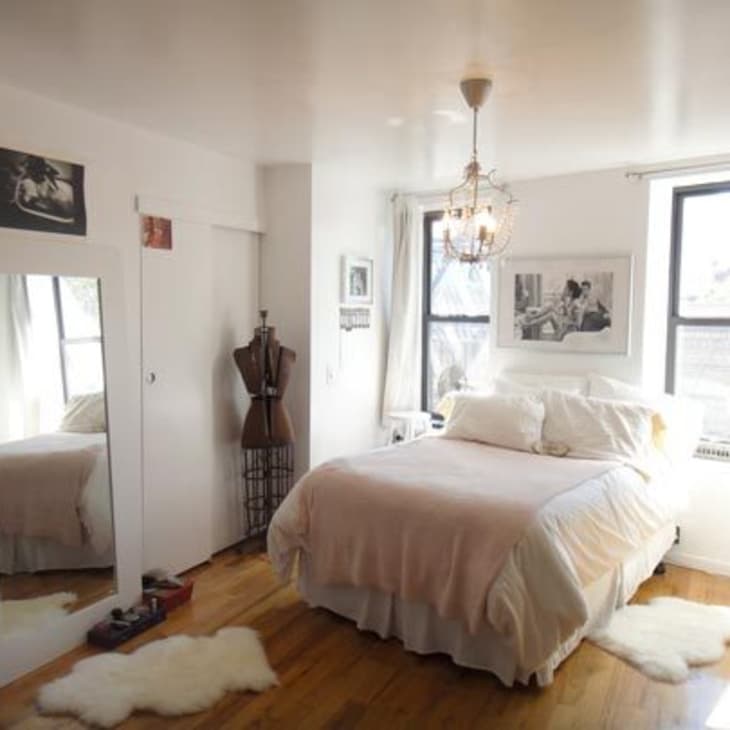 Cozy bedroom with a white bed, chandelier, dress form, books, and framed art on walls, featuring large windows.