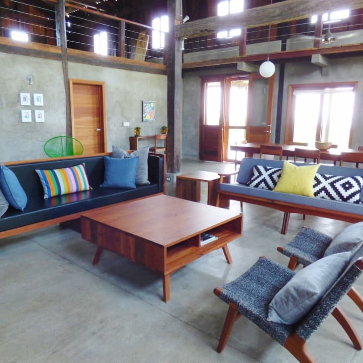 Spacious living room with wooden furniture, colorful cushions, and modern decor, featuring a loft and large windows.