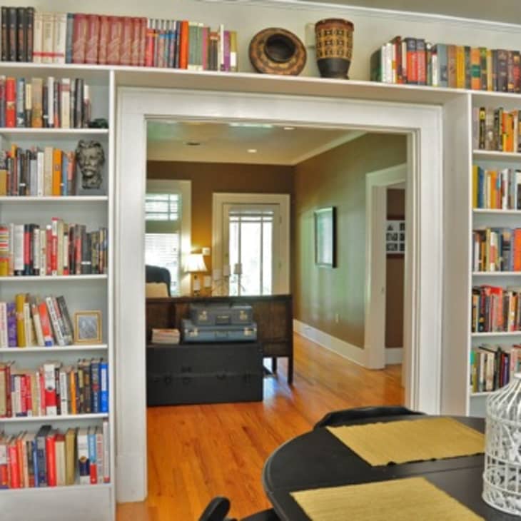 Home library with white bookshelves filled with books, leading to a cozy living room with a sofa and a window.