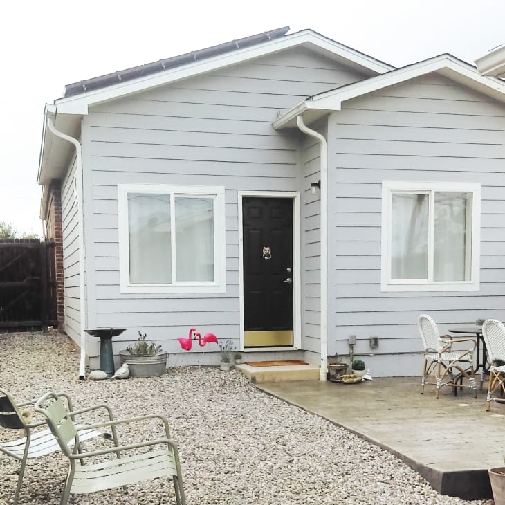 Small gray house with black door, gravel yard, metal chairs, pink flamingo decor, and outdoor dining set on patio.
