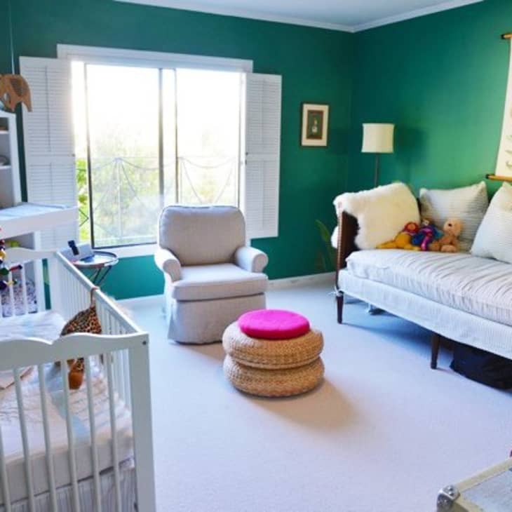 Nursery with green walls, crib, armchair, daybed, and botanical wall art, featuring a woven ottoman with a pink cushion.