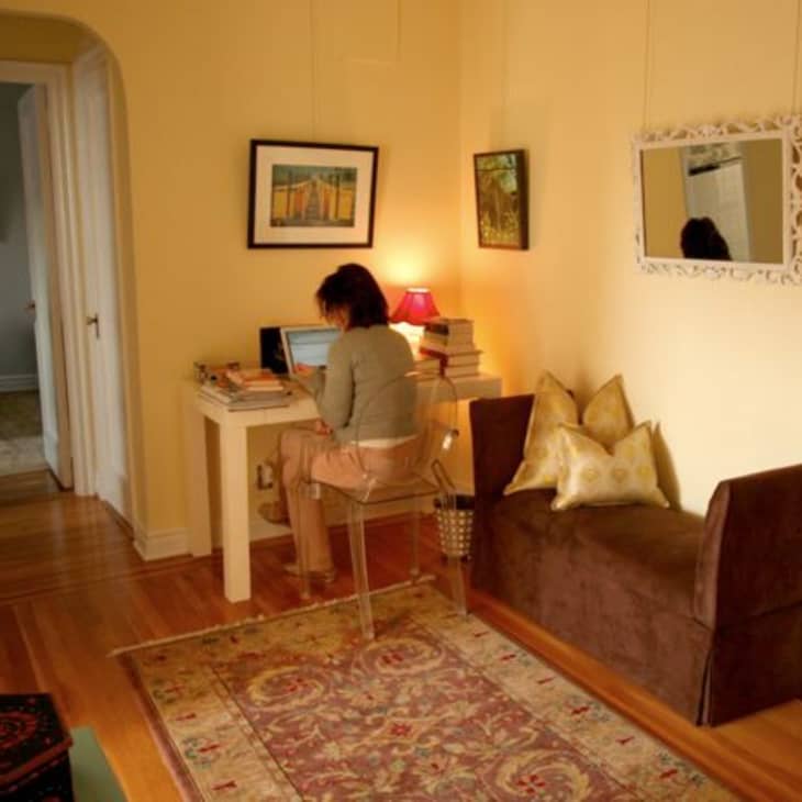 Woman working on a laptop at a white desk in a cozy room with a brown bench, decorative pillows, and framed art.