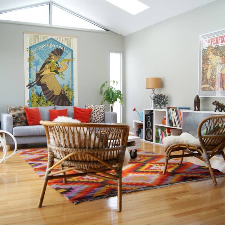 Living room with rattan chairs, colorful rug, gray sofa, and vibrant wall art. Bookshelves and decor items accent the space.