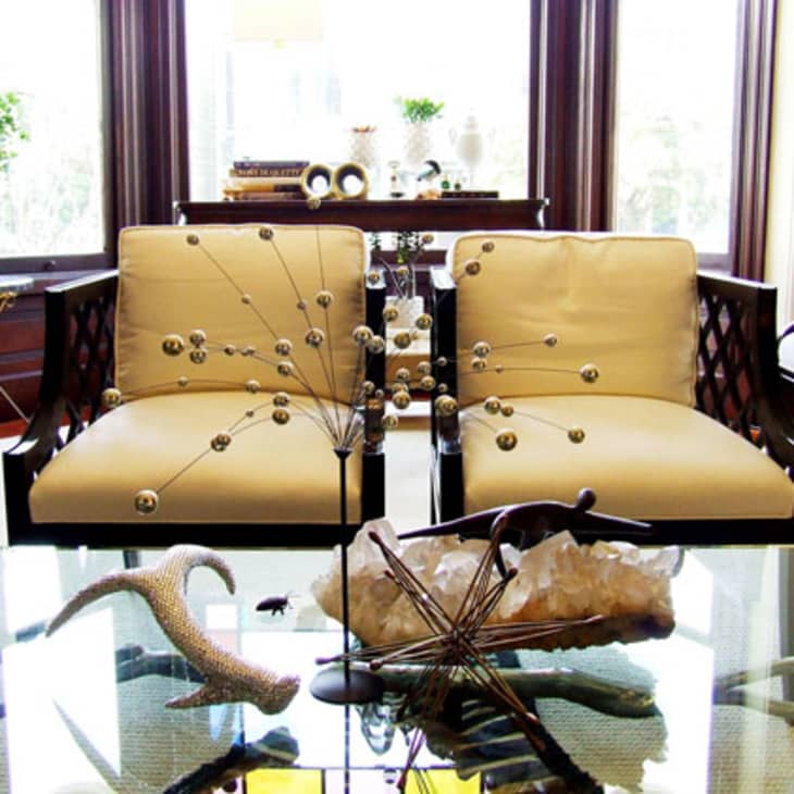 Living room with beige armchairs, glass coffee table, abstract decor, and large windows.