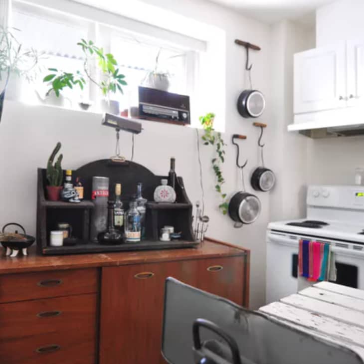 Small kitchen with white cabinets, hanging pots, a stove, and a wooden sideboard with plants and bottles.