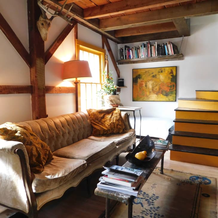 Cozy living room with a beige sofa, wooden beams, yellow stairs, bookshelves, and abstract art on the wall.