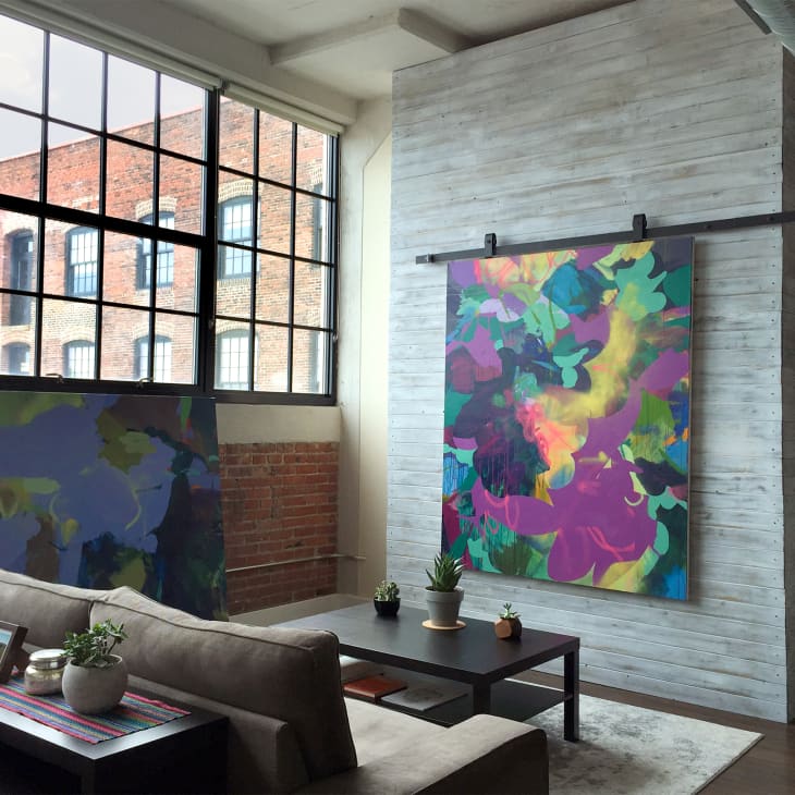 Loft living room with large abstract paintings, brick walls, and industrial windows. Sofa and coffee table with potted plants.