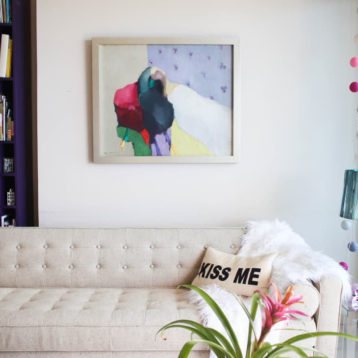 Beige tufted sofa with "Kiss Me" pillow, abstract painting, purple bookshelf, and teal lamp in a cozy living room.