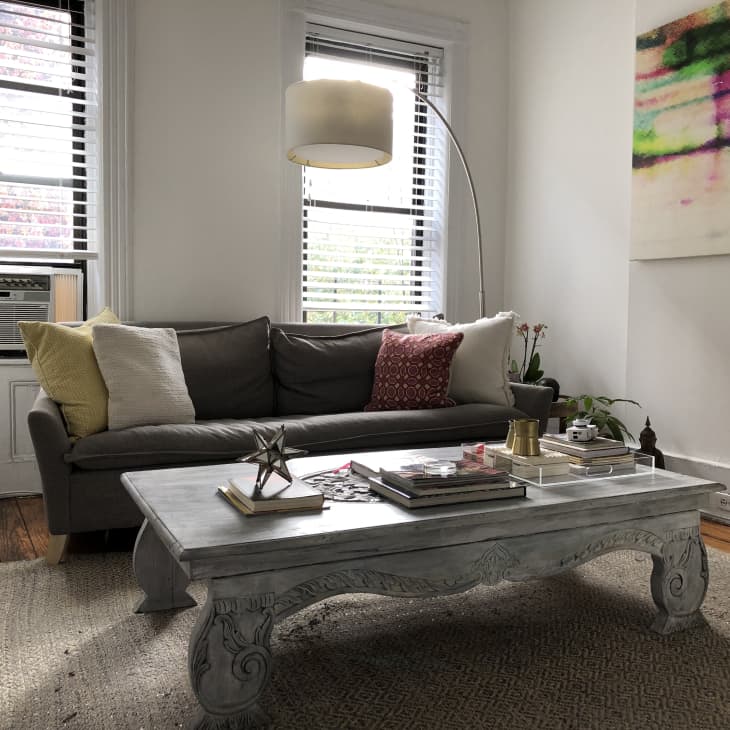 Living room with a gray sofa, colorful pillows, ornate coffee table, abstract wall art, and large windows with blinds.