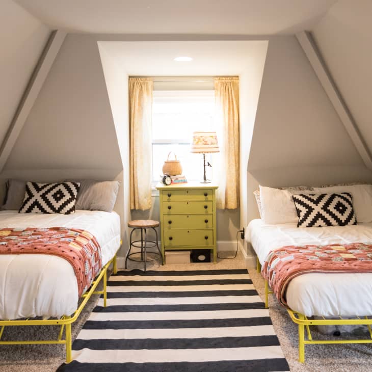 Attic bedroom with two beds, patterned pillows, colorful quilts, green nightstand, and striped rug.