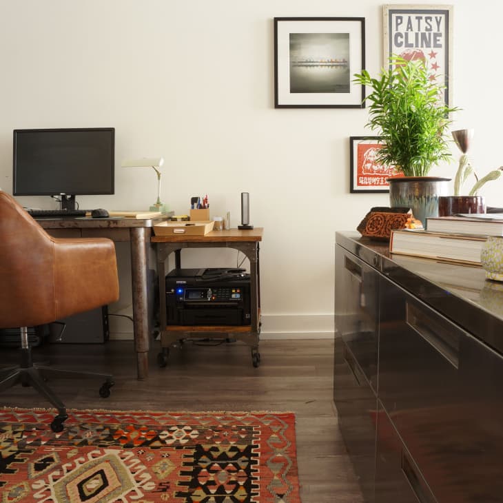 Home office with brown leather chair, wooden desk, computer, potted plant, framed art, and patterned rug.
