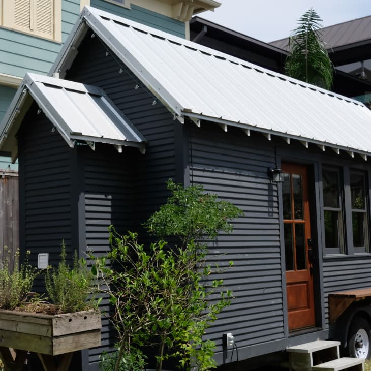 Dark gray tiny house on wheels with a white metal roof, wooden door, and small garden bed in front.