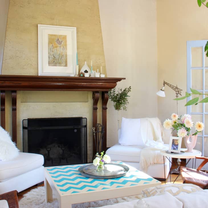 Cozy living room with white armchairs, a chevron-patterned coffee table, fireplace, and large windows with plants.