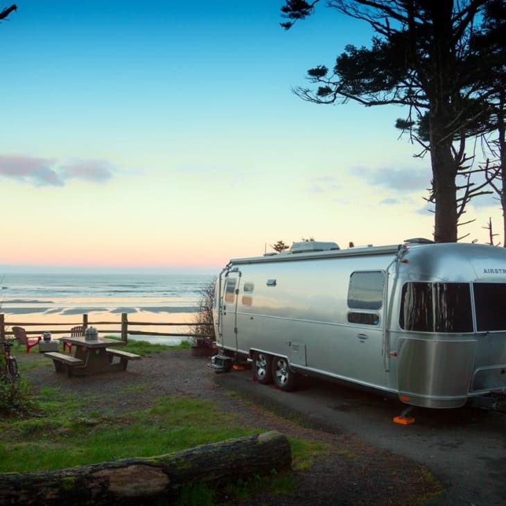 Airstream trailer parked at a coastal campsite with ocean view and picnic table at sunset.