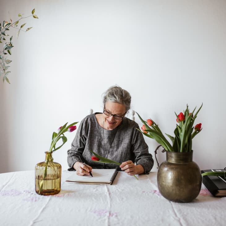 Woman writing at a table with tulips in vases and a plant in the background.