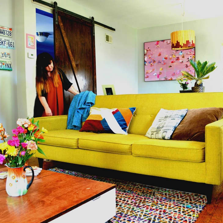 Living room with yellow sofa, colorful cushions, wall art, and a woman entering through a sliding barn door.