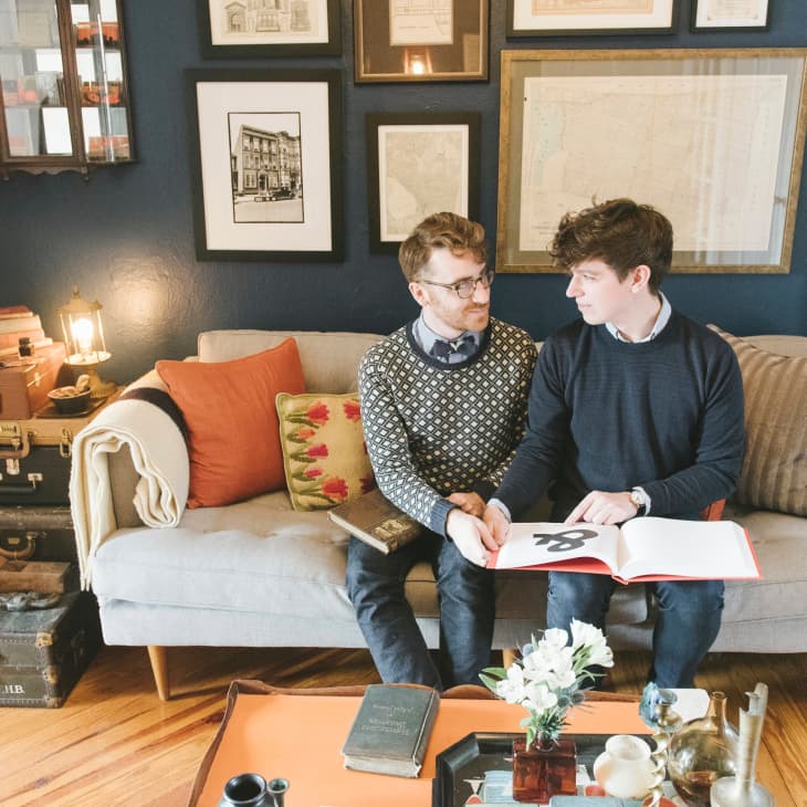 Two people sitting on a gray sofa in a cozy living room with framed art, books, and vintage suitcases.