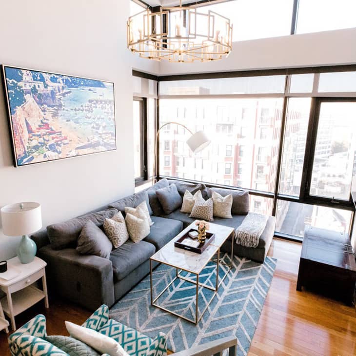 Modern living room with a gray sectional sofa, geometric rug, large windows, and a colorful wall painting.