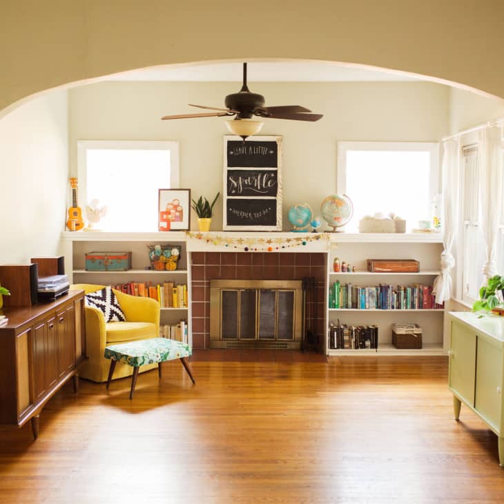 Living room with a yellow armchair, bookshelves, globes, and a fireplace, featuring a ceiling fan and wooden floors.
