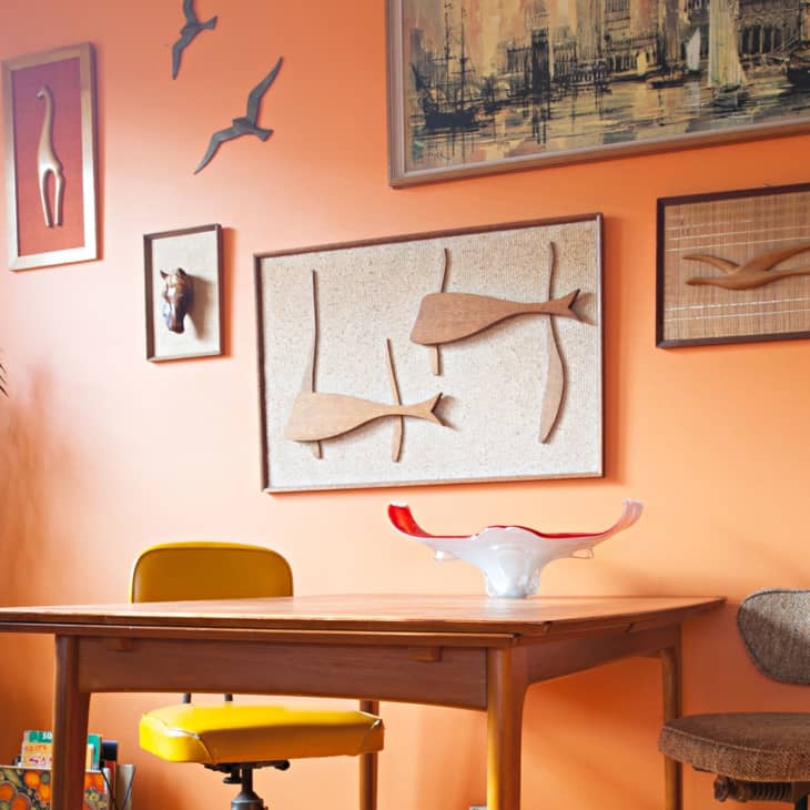 Mid-century modern dining area with orange walls, wooden table, yellow chair, and eclectic wall art.