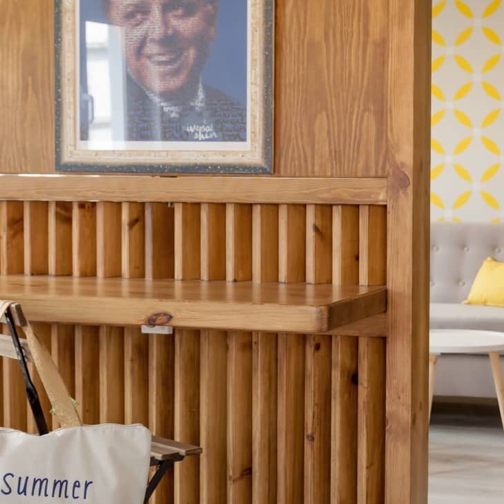 Wooden partition with a framed portrait, tote bag, and view of a gray sofa with a yellow pillow against patterned wallpaper.