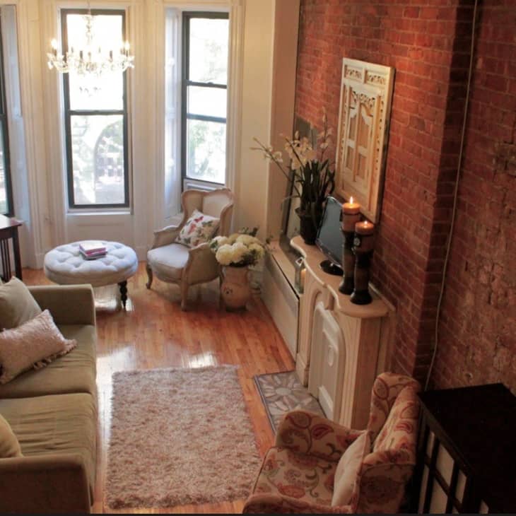 Cozy living room with a green sofa, brick wall, chandelier, and a desk by three large windows.