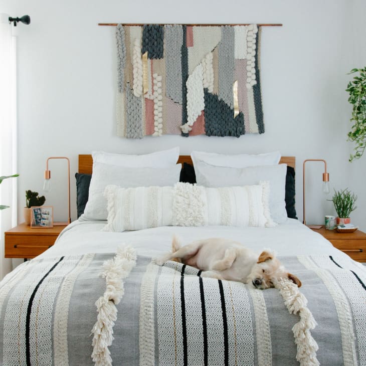A white bed with a striped blanket at the foot and a macrame wall hanging.
