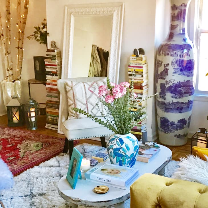 Cozy living room with a large vase, books, a mirror, floral decor, and a yellow sofa on a patterned rug.
