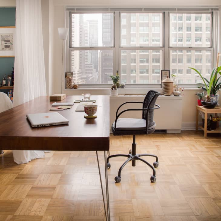 Home office with a wooden desk, laptop, swivel chair, and large window. Sofa with cushions and plants on the side.