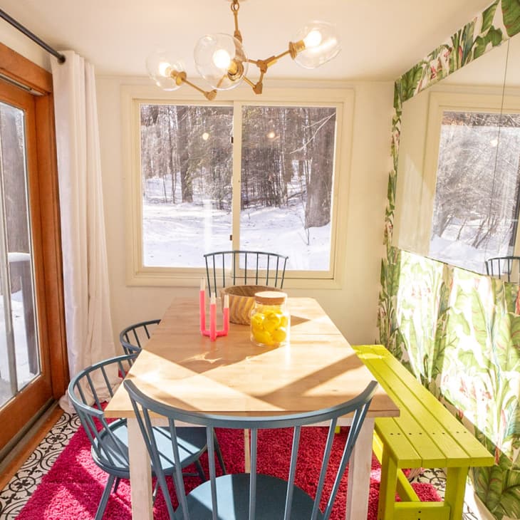 Dining room with a wooden table, blue chairs, yellow bench, and tropical wallpaper, overlooking a snowy landscape.