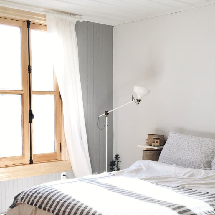 Cozy bedroom with a wooden window, white curtains, patterned bedding, wall lamp, and small plant on a side table.