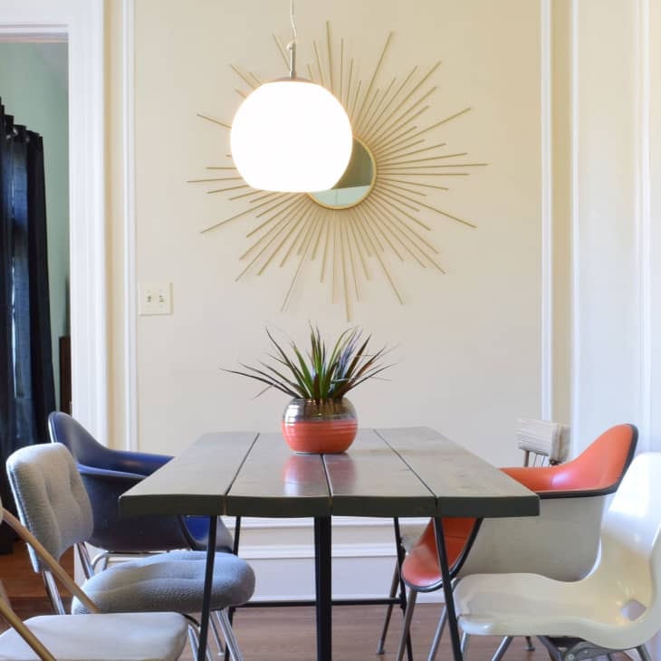 Dining room with a wooden table, mixed chairs, a potted plant centerpiece, and a sunburst mirror on the wall.