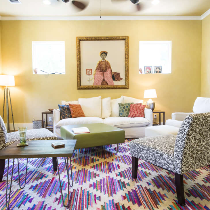 Living room with patterned chairs, colorful rug, white sofa, and framed artwork on yellow walls. Bookshelf and lamps add decor.