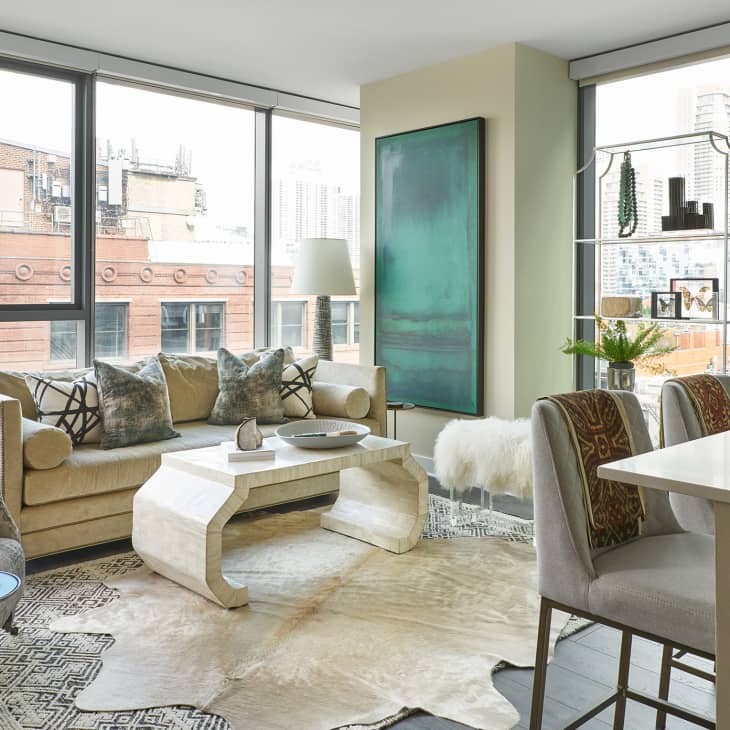 Modern living room with large windows, beige sofa, abstract painting, and decorative pillows, overlooking city buildings.