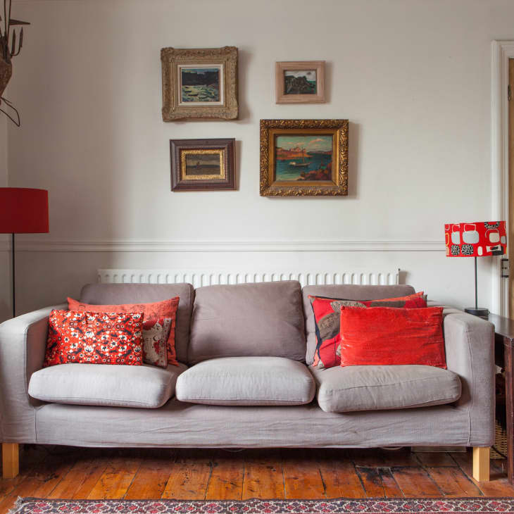 Gray sofa with red and patterned cushions, framed art on wall, wooden floor, and red lampshade in cozy living room.