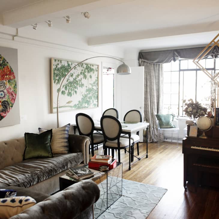 Living room with a gray sofa, colorful wall art, dining table, piano, and large window with curtains.