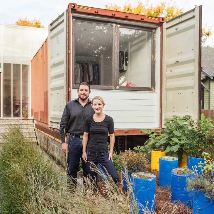 Couple standing in front of a shipping container home with a garden of blue planters.