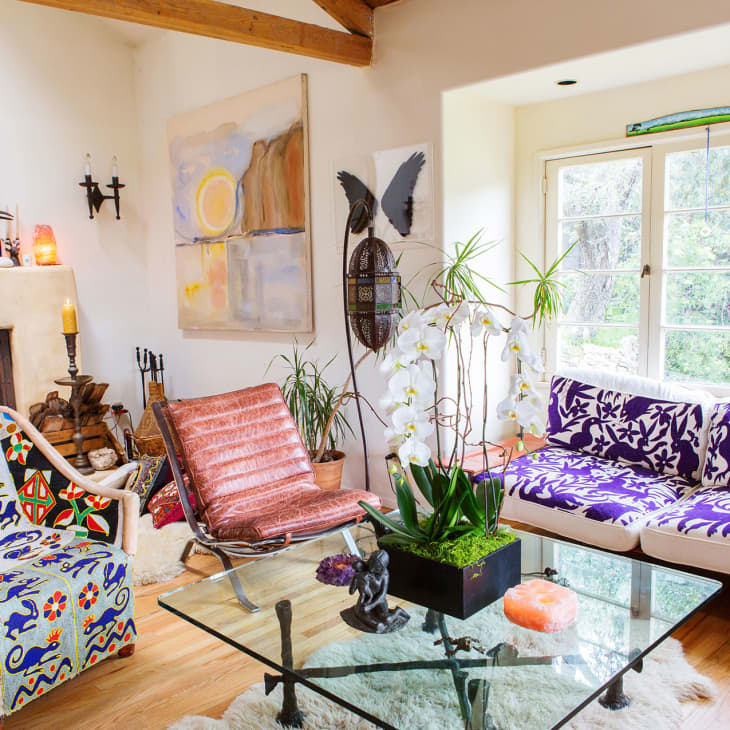 Eclectic living room with colorful armchair, purple floral sofa, glass coffee table, and large windows.