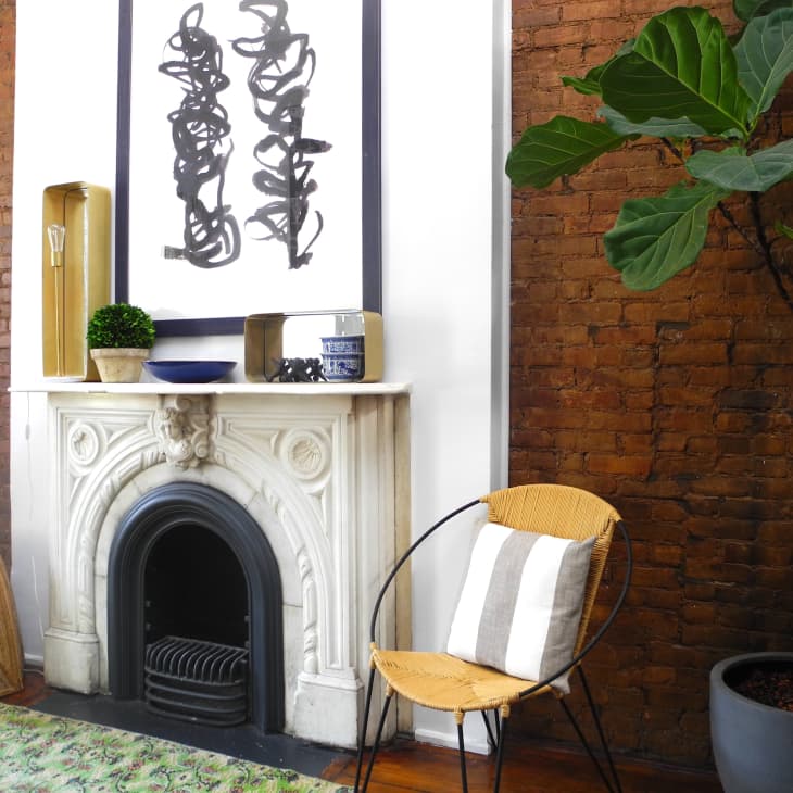 Brick-walled living room with a white fireplace, abstract art, wicker chair, striped pillow, and a potted fiddle leaf fig tree.