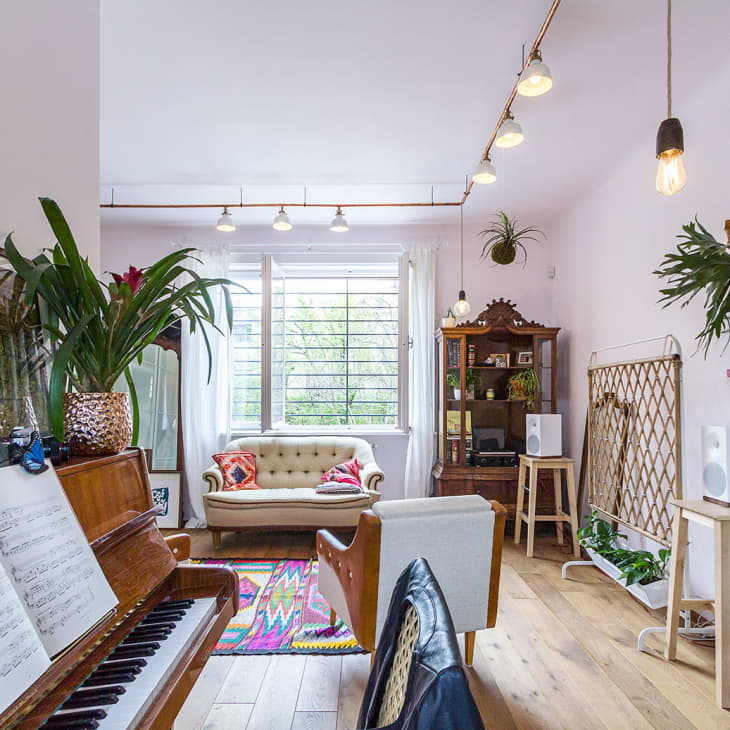 Living room with piano, sheet music, plants, beige sofa, colorful rug, and vintage cabinet near large window.