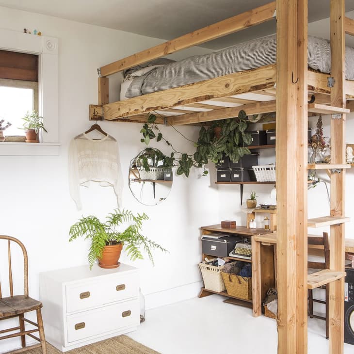 Loft bed with wooden ladder, plants, and storage underneath in a cozy room with a chair and white drawers.