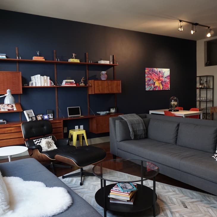 Modern living room with dark blue accent wall, gray sofas, wooden shelving, and colorful floral artwork above a dining table.