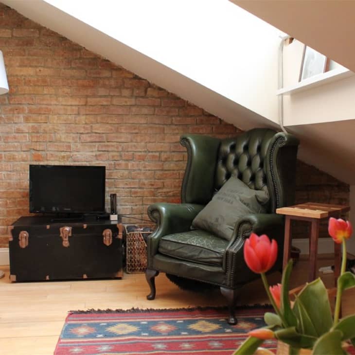 Attic room with a green leather armchair, brick wall, TV on a trunk, floor lamp, and red tulips in the foreground.