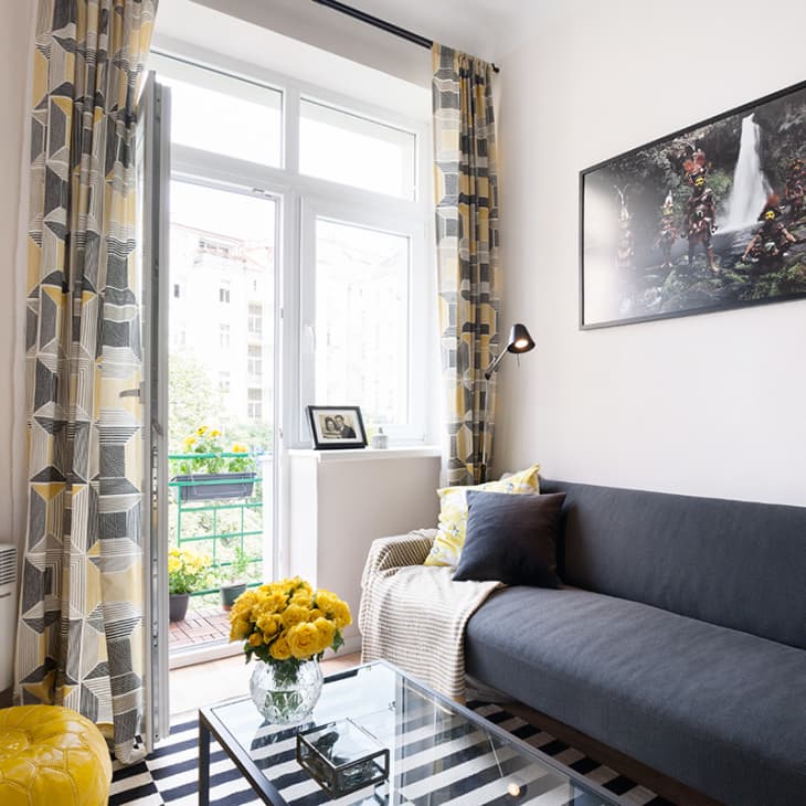 Living room with gray sofa, geometric curtains, yellow ottoman, and glass coffee table with yellow flowers.
