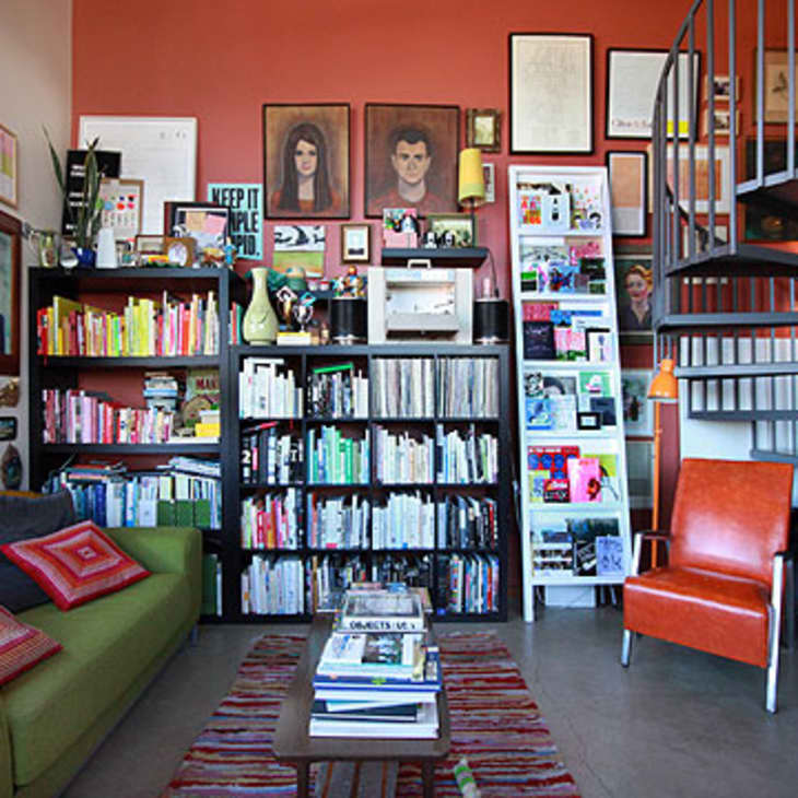 Eclectic living room with green sofa, colorful bookshelves, spiral staircase, and framed portraits on orange wall.