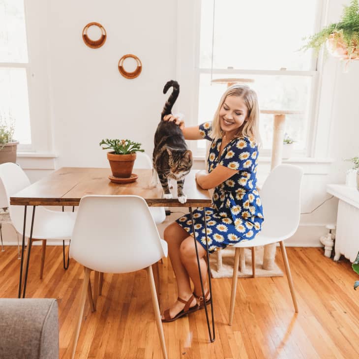 Woman in a floral dress petting a cat on a wooden dining table surrounded by plants and white chairs.